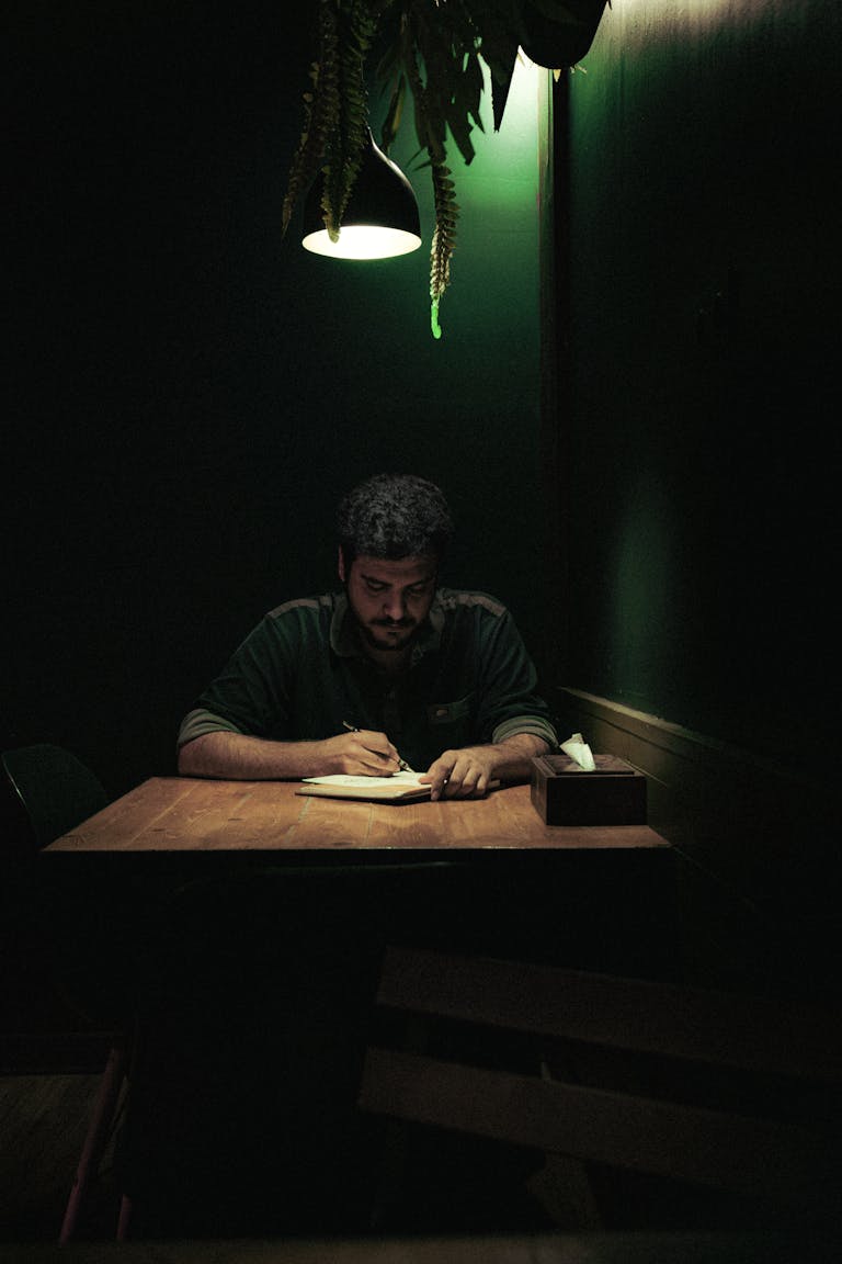 A man writes under a dim green light creating a contemplative atmosphere.